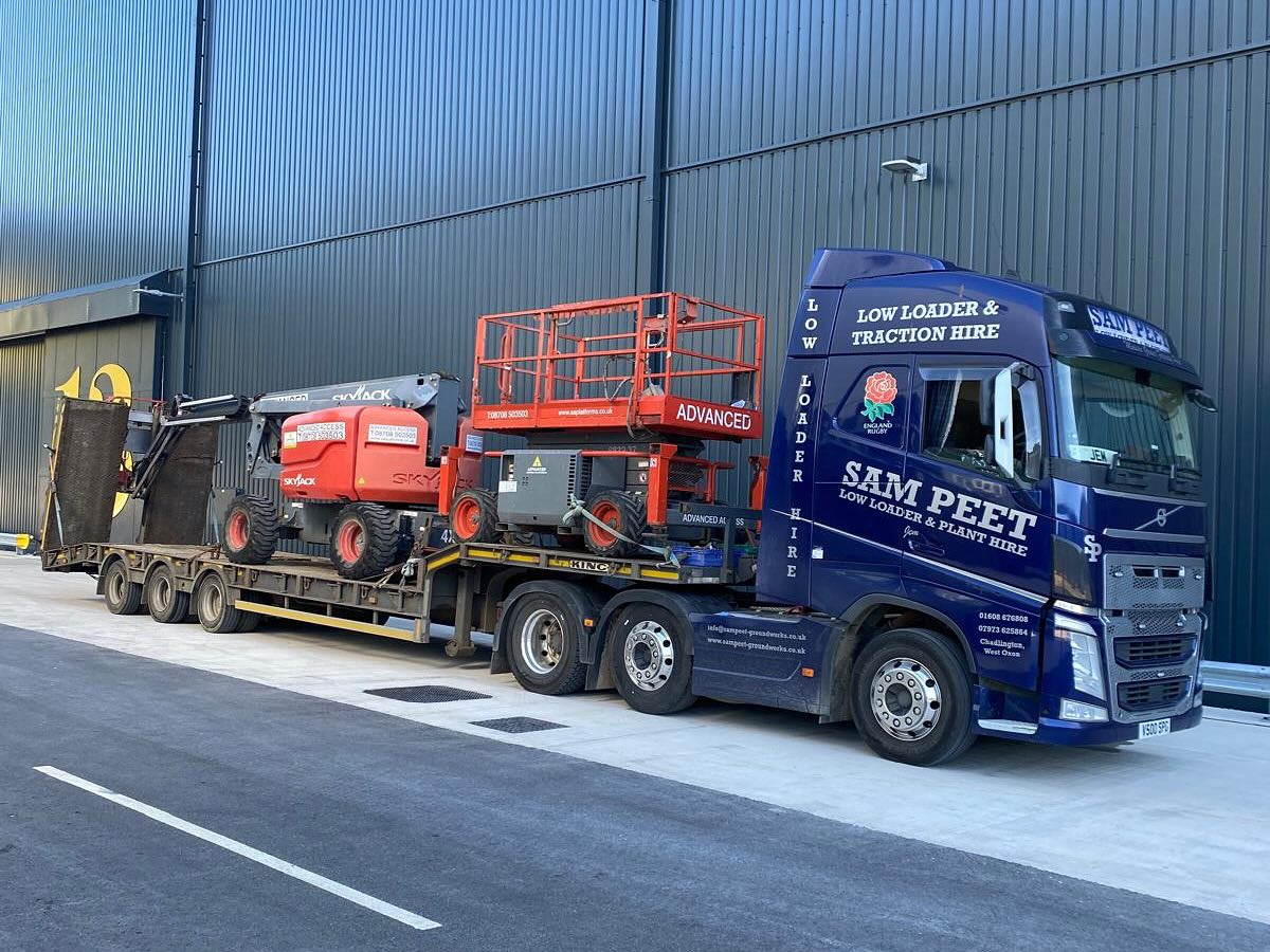 A blue SAM PEET truck hauls a flatbed trailer carrying industrial equipment, including a red cherry picker and a scissor lift, parked beside a large industrial building.