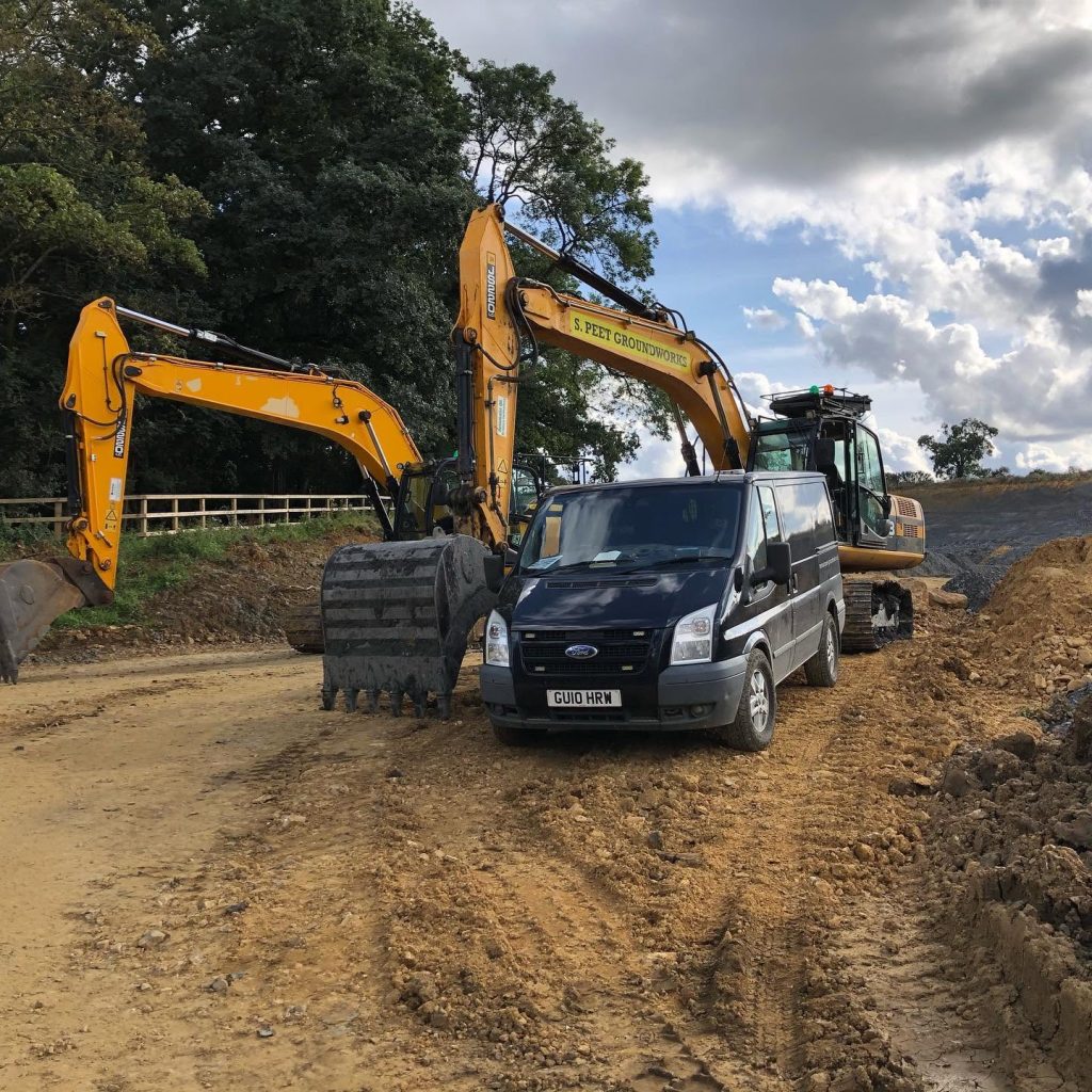A black van is parked on a dirt construction site between two large yellow excavators, with trees and clouds visible in the background.