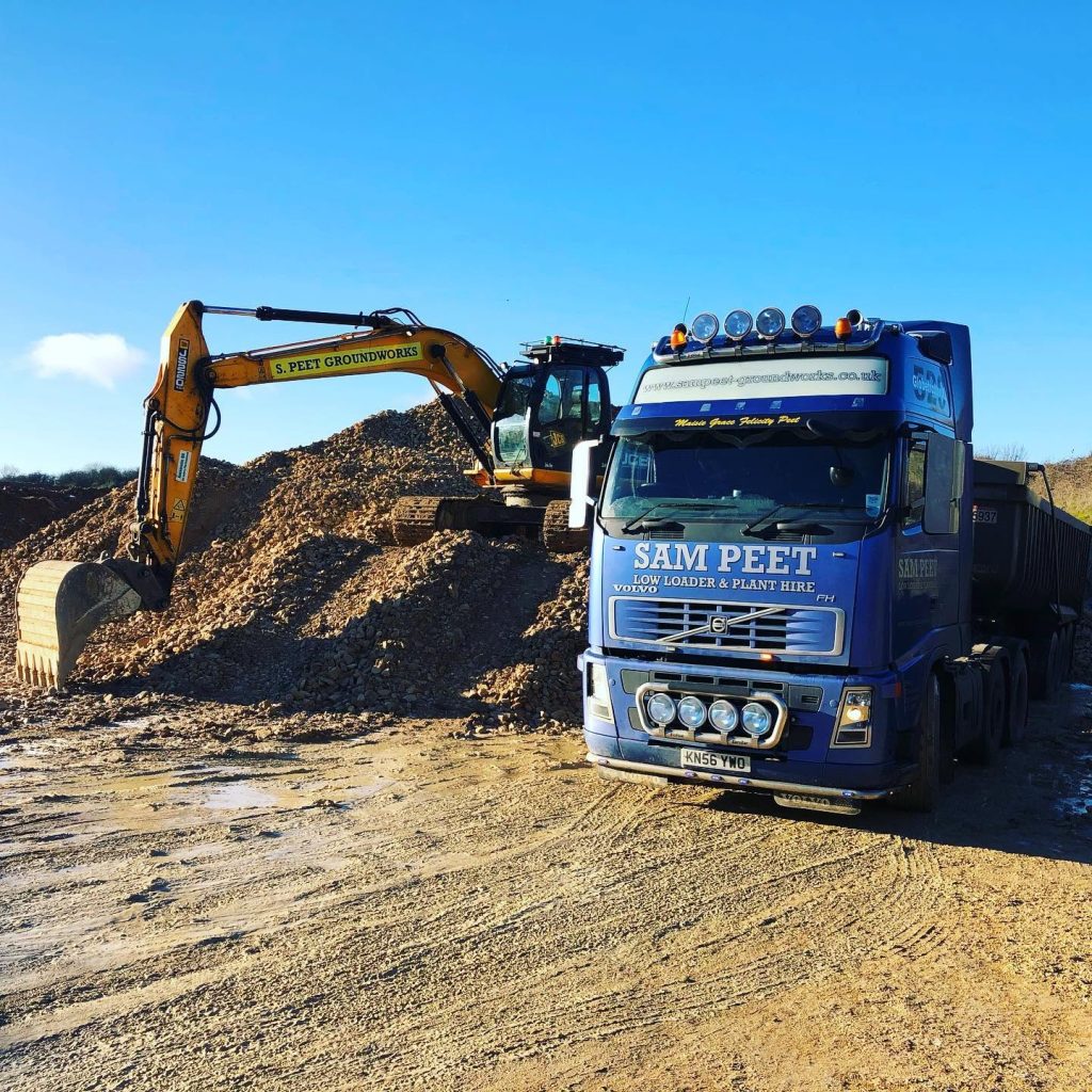 A blue Sam Peet truck is parked on a construction site next to an orange excavator digging into a pile of gravel under a sunny sky.