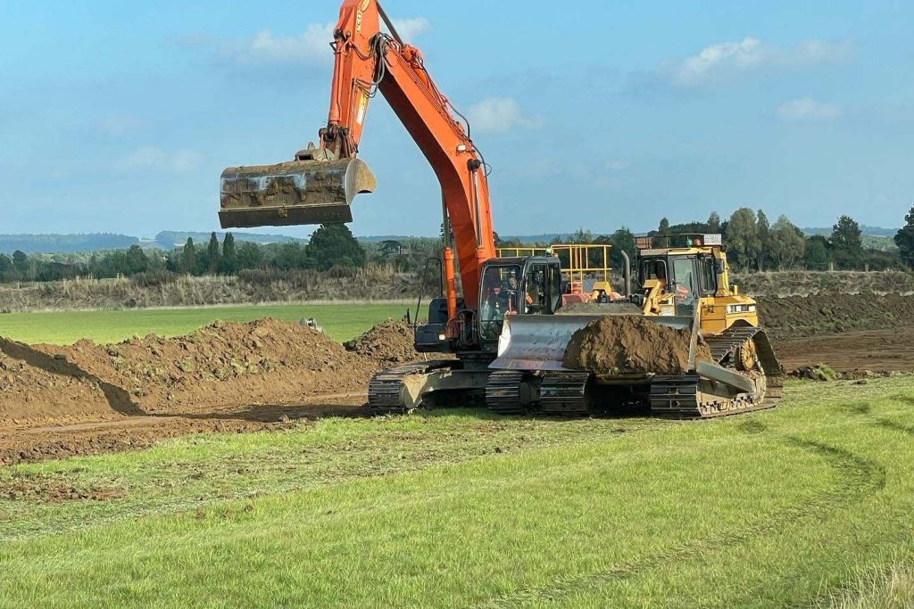 An orange excavator and two bulldozers are working on a construction site, moving earth in a grassy field with trees and blue sky in the background.