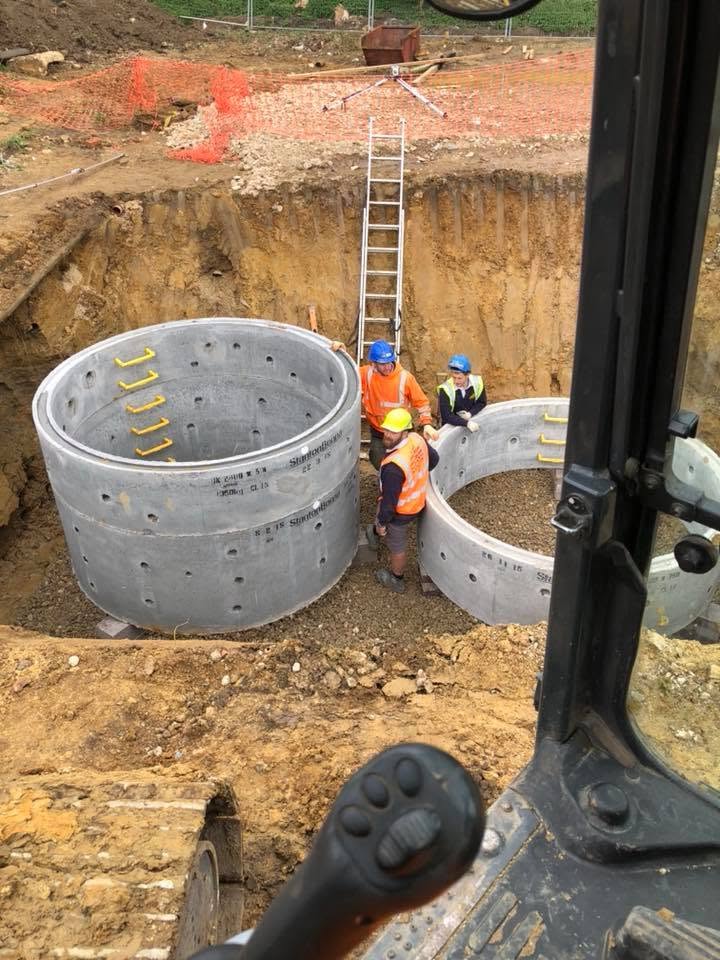 Three construction workers stand inside a deep excavation site beside two large concrete culvert sections. A metal ladder leans against the dirt wall, and construction equipment is visible in the foreground.