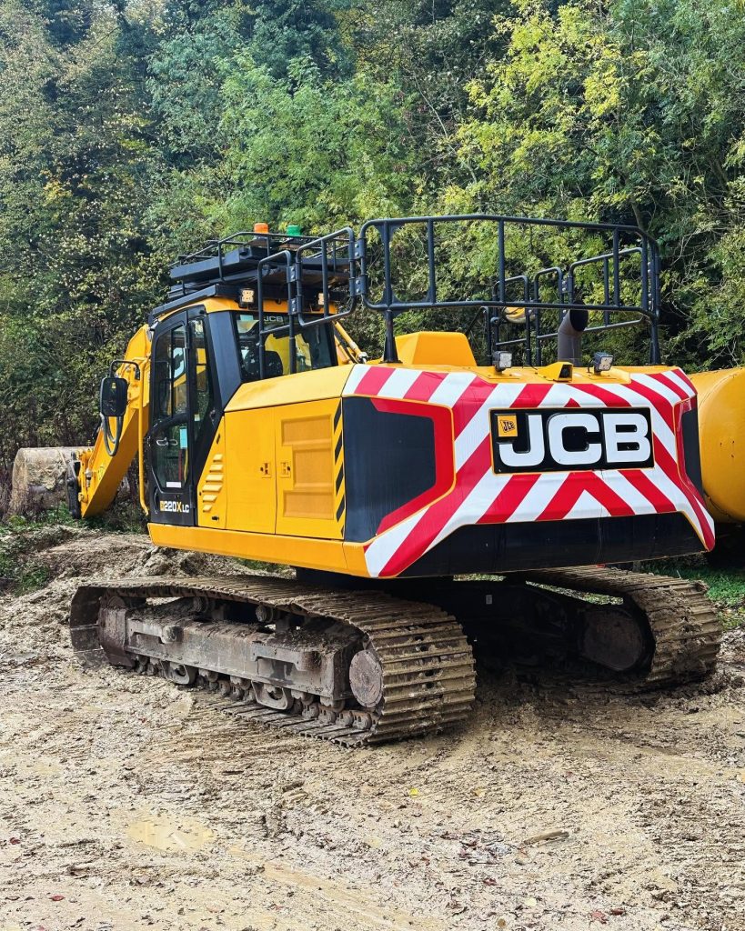 A yellow JCB excavator with red and white stripes on the back is parked on a muddy construction site, surrounded by trees and greenery.