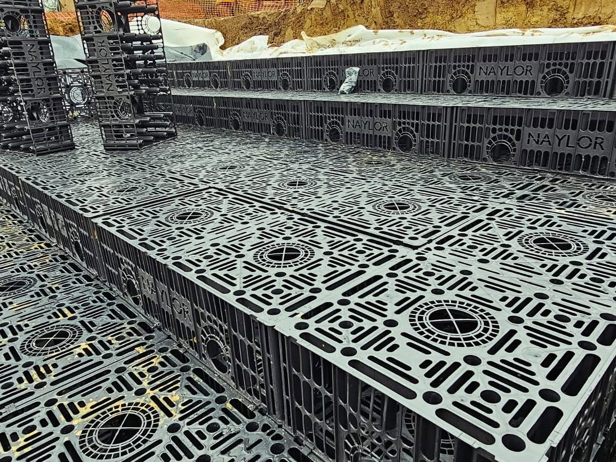 Plastic modular crates are stacked and arranged at a construction site, forming a patterned surface. The crates are labeled “Naylor” and used for stormwater management or drainage systems. Soil and construction materials are visible in the background.