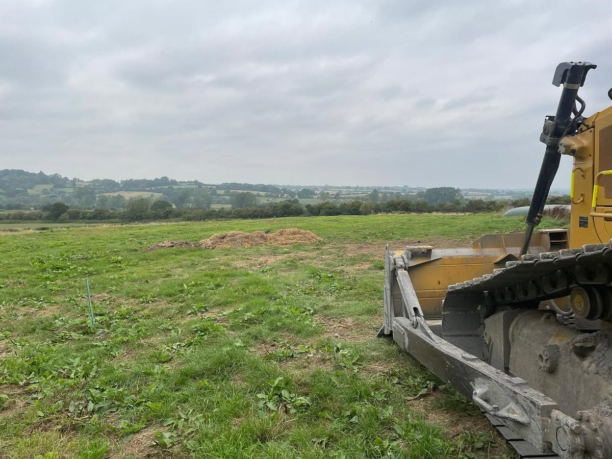 A bulldozer is parked on a grassy field under a cloudy sky, with hills and trees visible in the distance.