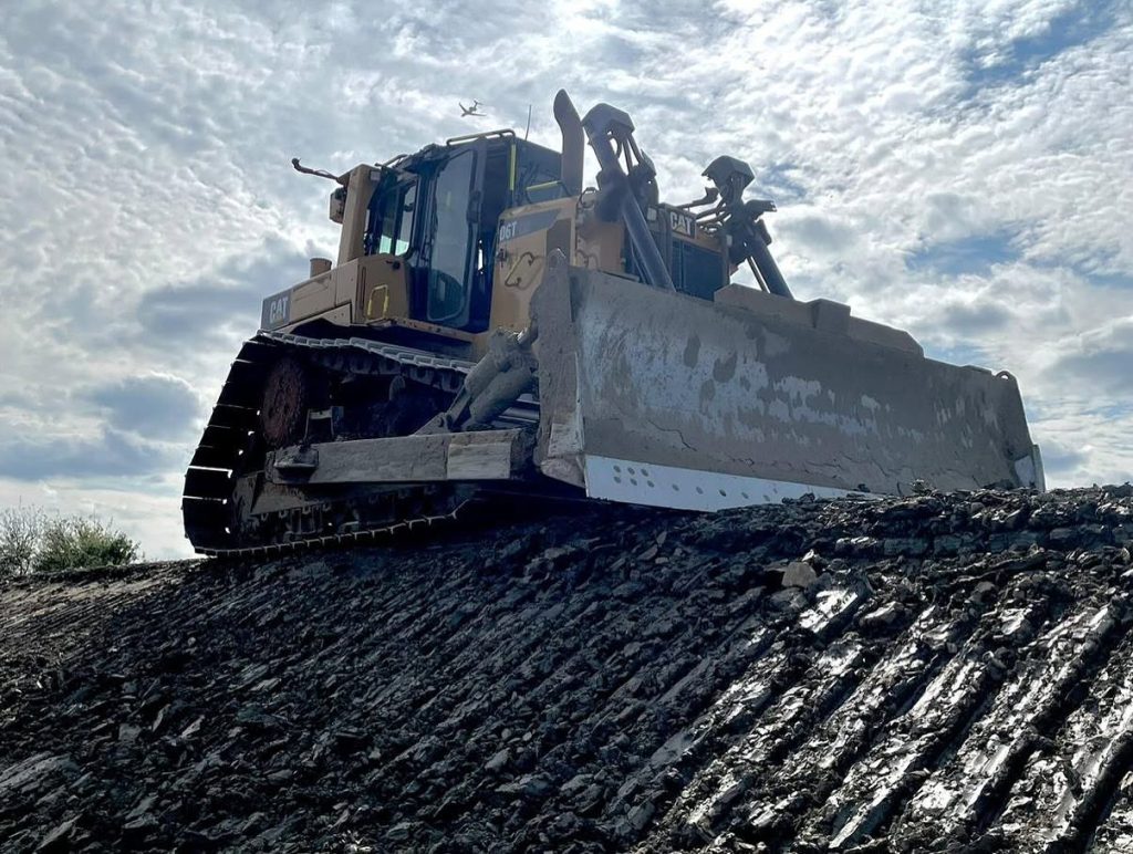 A large bulldozer pushes soil up a muddy, uneven hill under a cloudy sky, with sunlight breaking through the clouds.