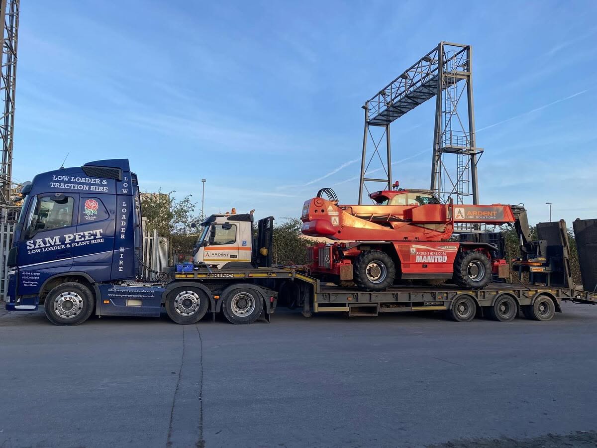 A blue SAM PEET truck hauls a flatbed trailer carrying a large red Manitou telehandler and another yellow truck in an industrial area under a blue sky.