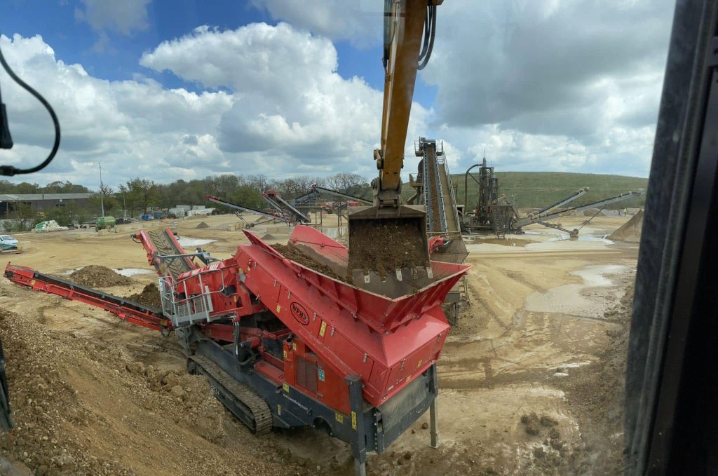 A large excavator drops dirt into a red industrial screening machine at a construction site, with sandy ground, equipment, and cloudy skies in the background.