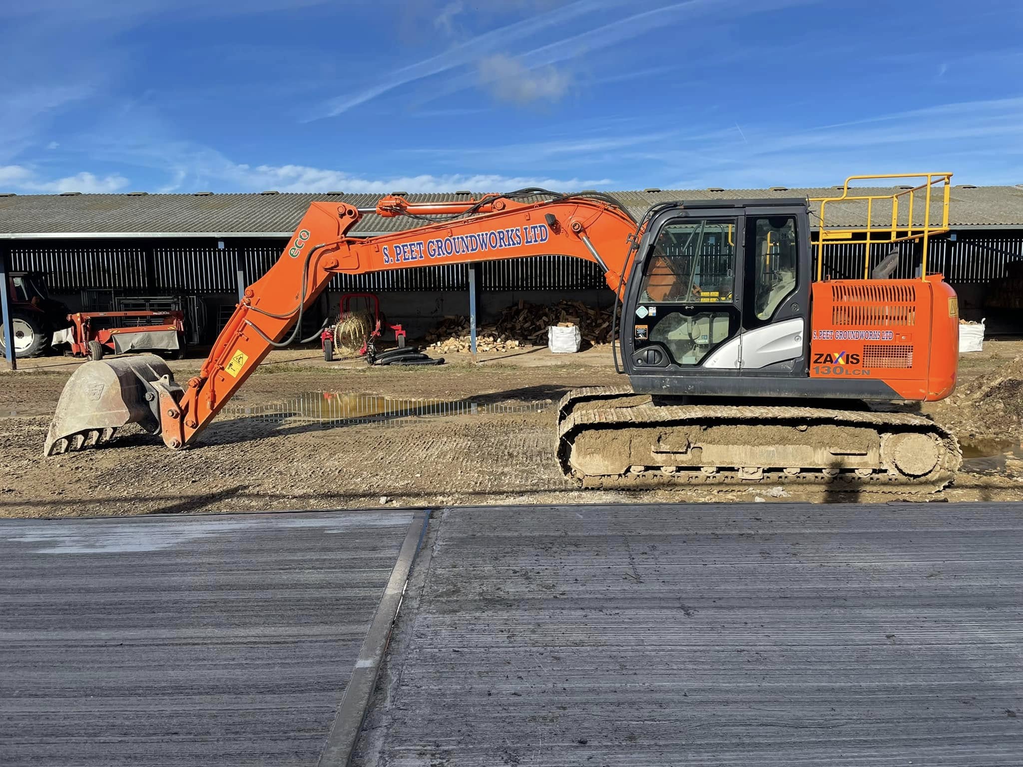 An orange excavator labeled “3 Feet Groundworks Ltd” is parked on a construction site next to a building. The sky is clear and blue.