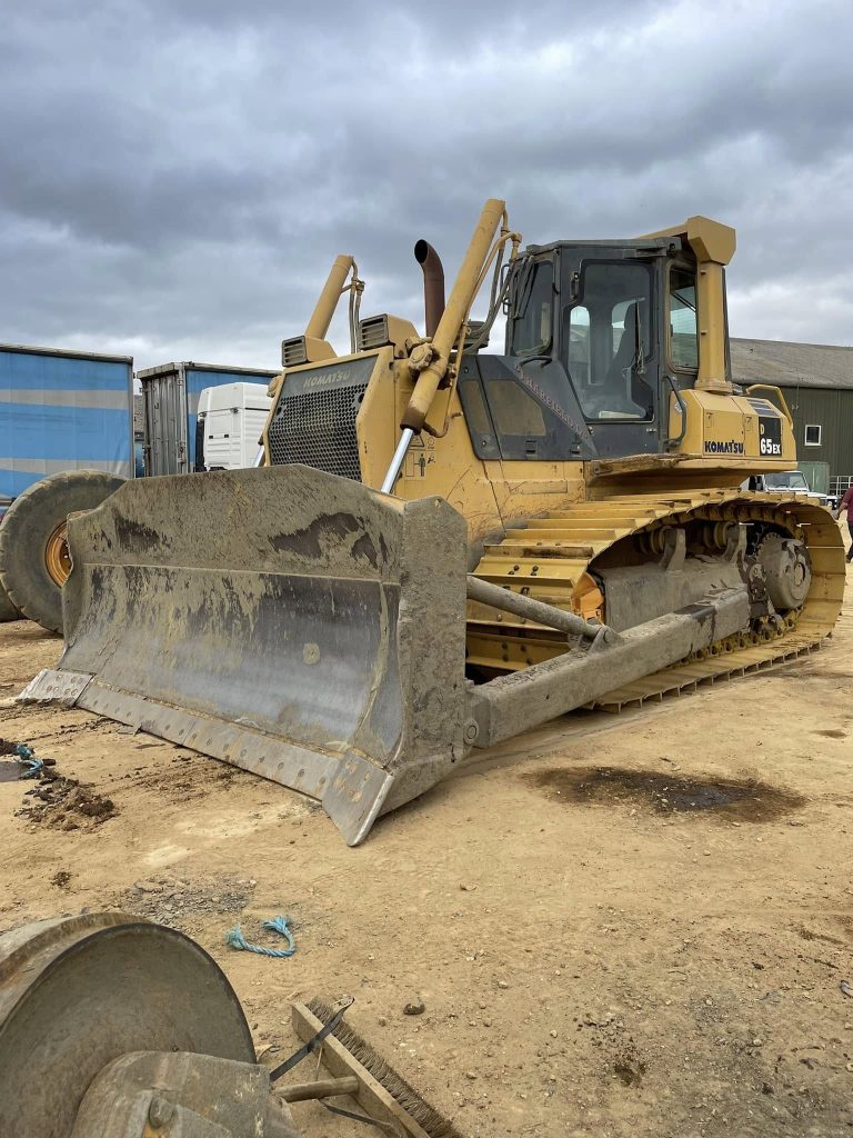 A large yellow bulldozer with a muddy blade is parked on a construction site, surrounded by dirt, equipment, and buildings under a cloudy sky.