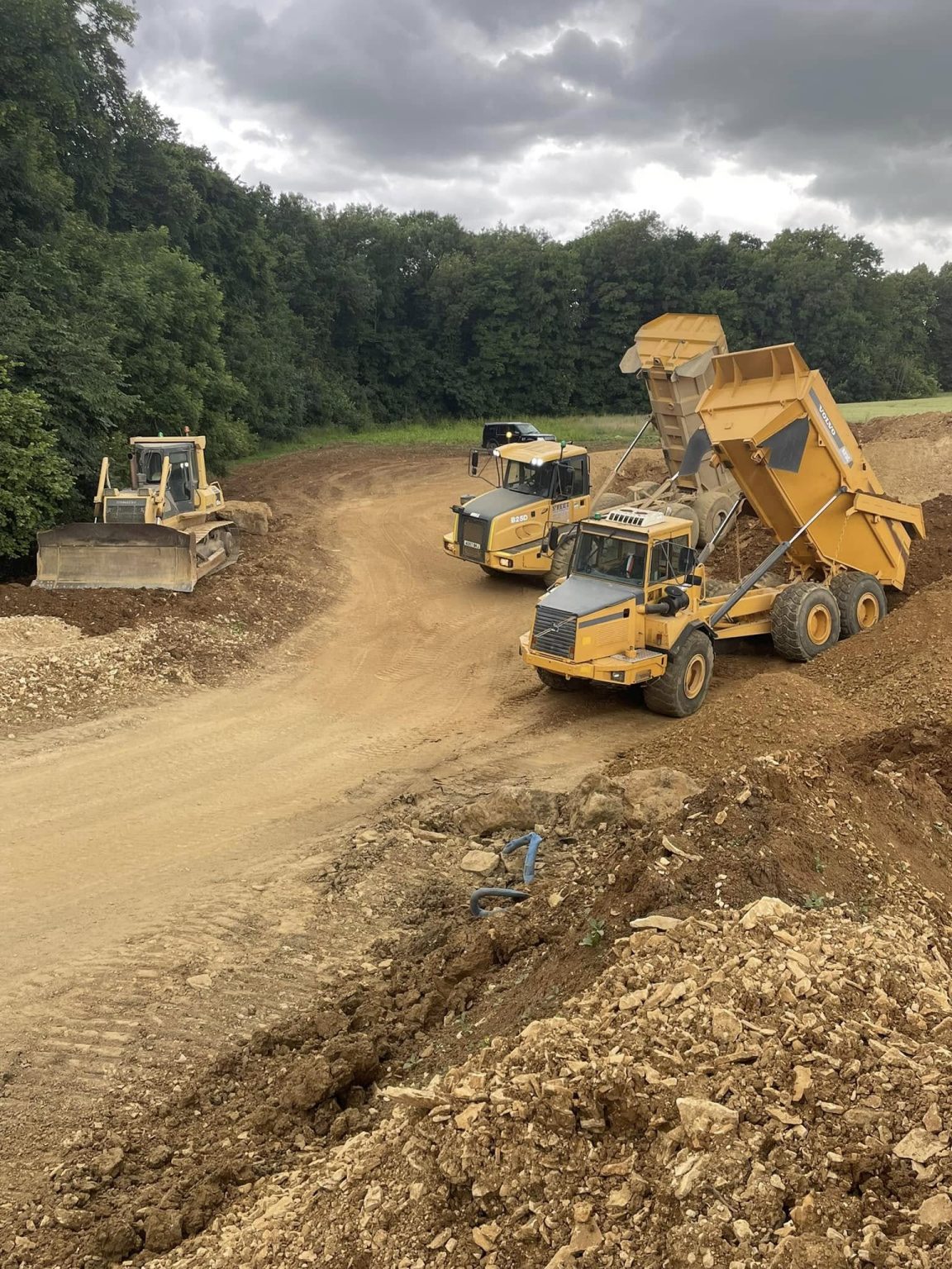 Three large construction vehicles, including a bulldozer and a dump truck unloading dirt, work on a dirt road at a construction site surrounded by trees under a cloudy sky.