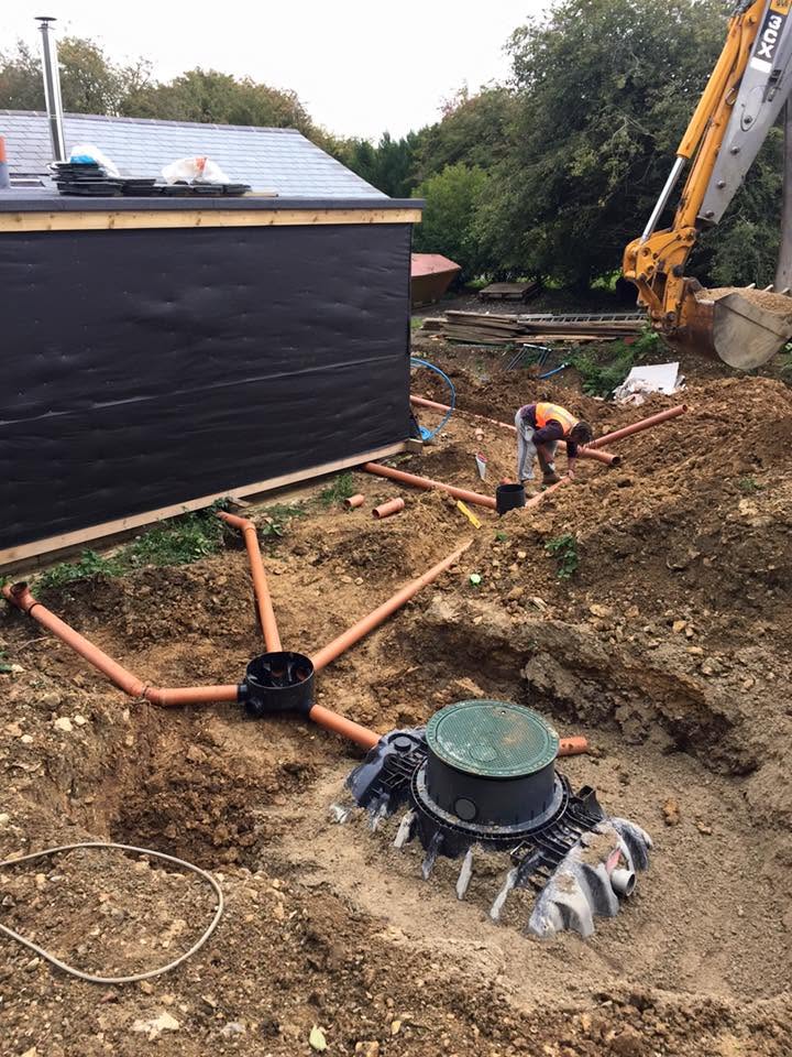 A construction worker connects orange pipes to a black building and a large green tank, with a yellow excavator nearby and dirt piles around the site.