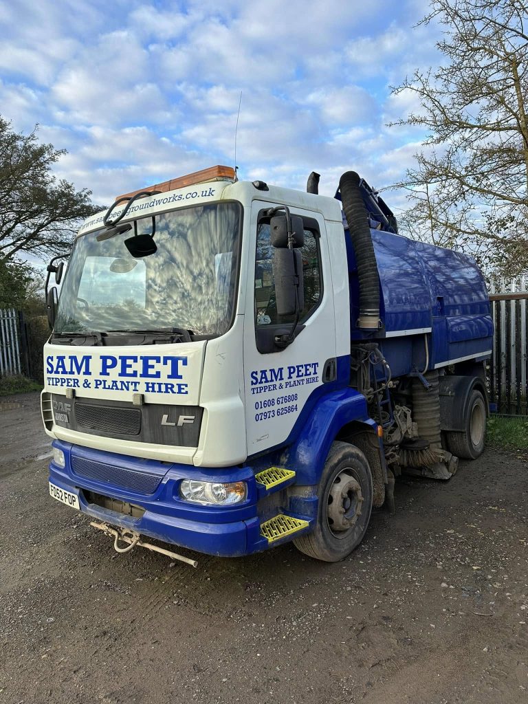 A blue and white vacuum truck labeled “SAM PEET TIPPER & PLANT HIRE” is parked on a gravel surface near a fence and trees under a partly cloudy sky.
