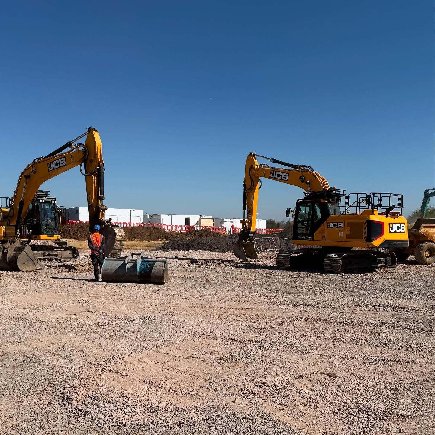 A construction site with two yellow JCB excavators on gravel, a worker in an orange vest standing nearby, and industrial buildings in the background under a clear blue sky.