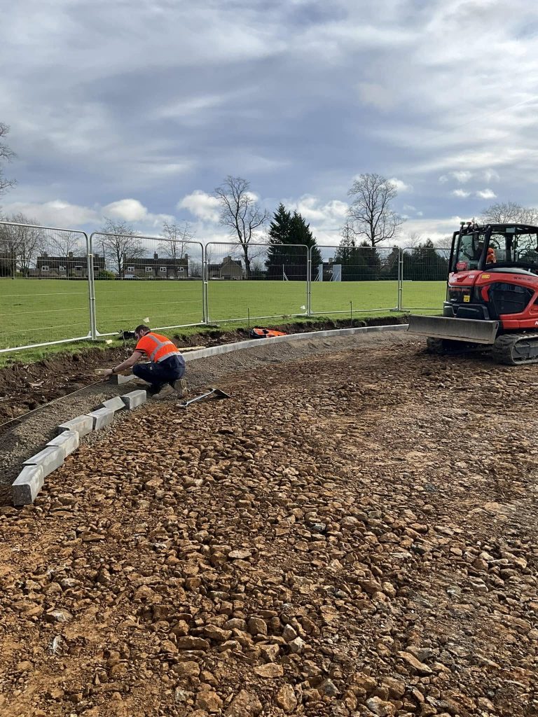 A worker in a high-visibility vest installs curb stones along a gravel path, while another person operates an excavator nearby. Metal fencing and a grassy field with trees are visible in the background under a cloudy sky.