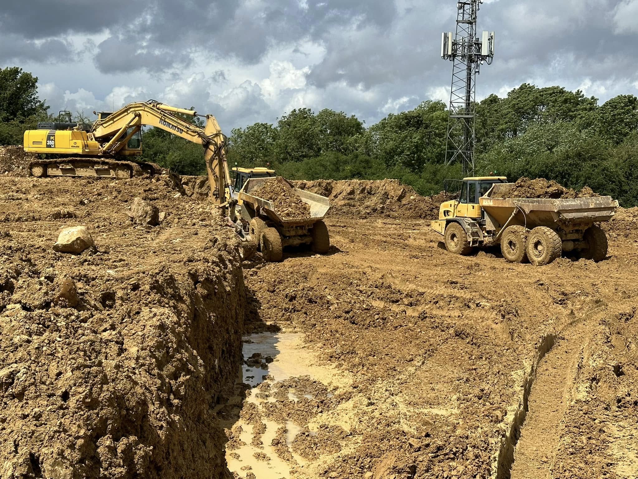 Two large yellow construction vehicles work on a muddy site, with one excavator loading dirt into a dump truck. Another dump truck waits nearby. Trees and a cell tower are visible in the background under a cloudy sky.