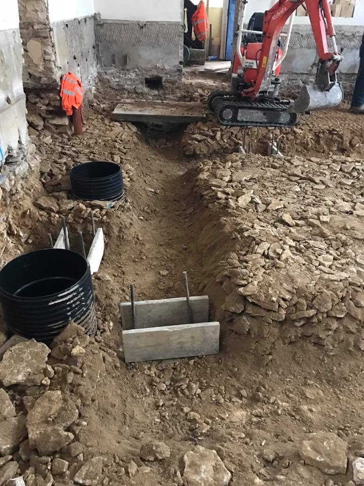 A construction site with exposed dirt, trenches, two black plastic cylinders, a small concrete form, and a mini-excavator. Partially demolished walls and workers in orange vests are visible in the background.