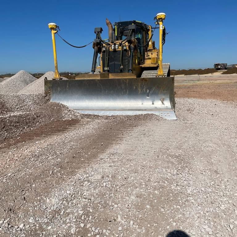 A bulldozer is pushing a pile of gravel on a construction site under a clear blue sky. The ground is covered with small rocks, and mounds of gravel are nearby.