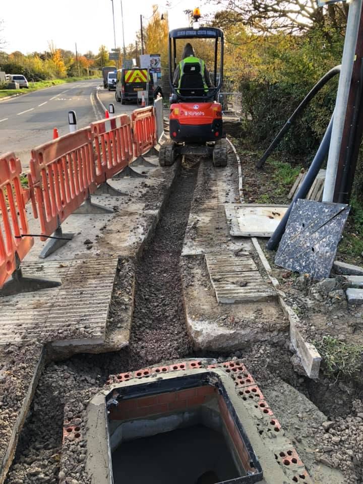 A construction worker operates a small excavator, digging a narrow trench along a roadside. Orange barriers line the sidewalk, and a utility box is partially installed at the front.