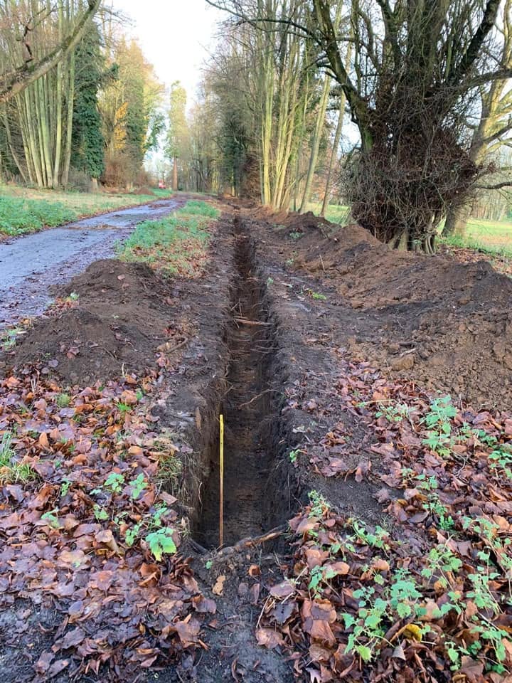 A long, narrow trench is dug beside a rural path lined with bare trees. The ground is covered with fallen leaves and some green plants. A yellow marker is visible inside the trench.