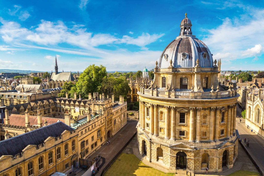 Aerial view of the Radcliffe Camera, a domed library building at Oxford University, England, surrounded by historic stone buildings and a clear blue sky.
