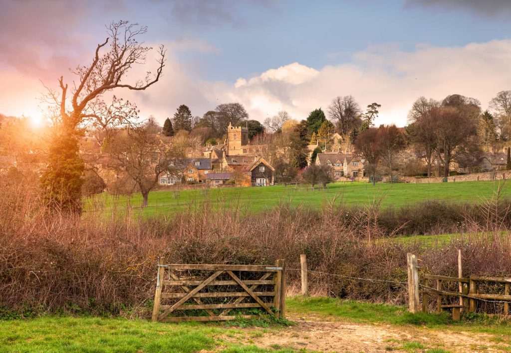 A wooden gate opens to a grassy field with a village and church tower in the background, surrounded by trees under a partly cloudy sky at sunset.