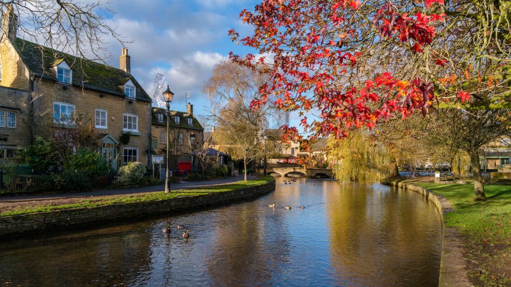 A calm river flows through a village with stone houses on the left, autumn trees with red leaves on the right, and ducks swimming in the water under a blue sky.