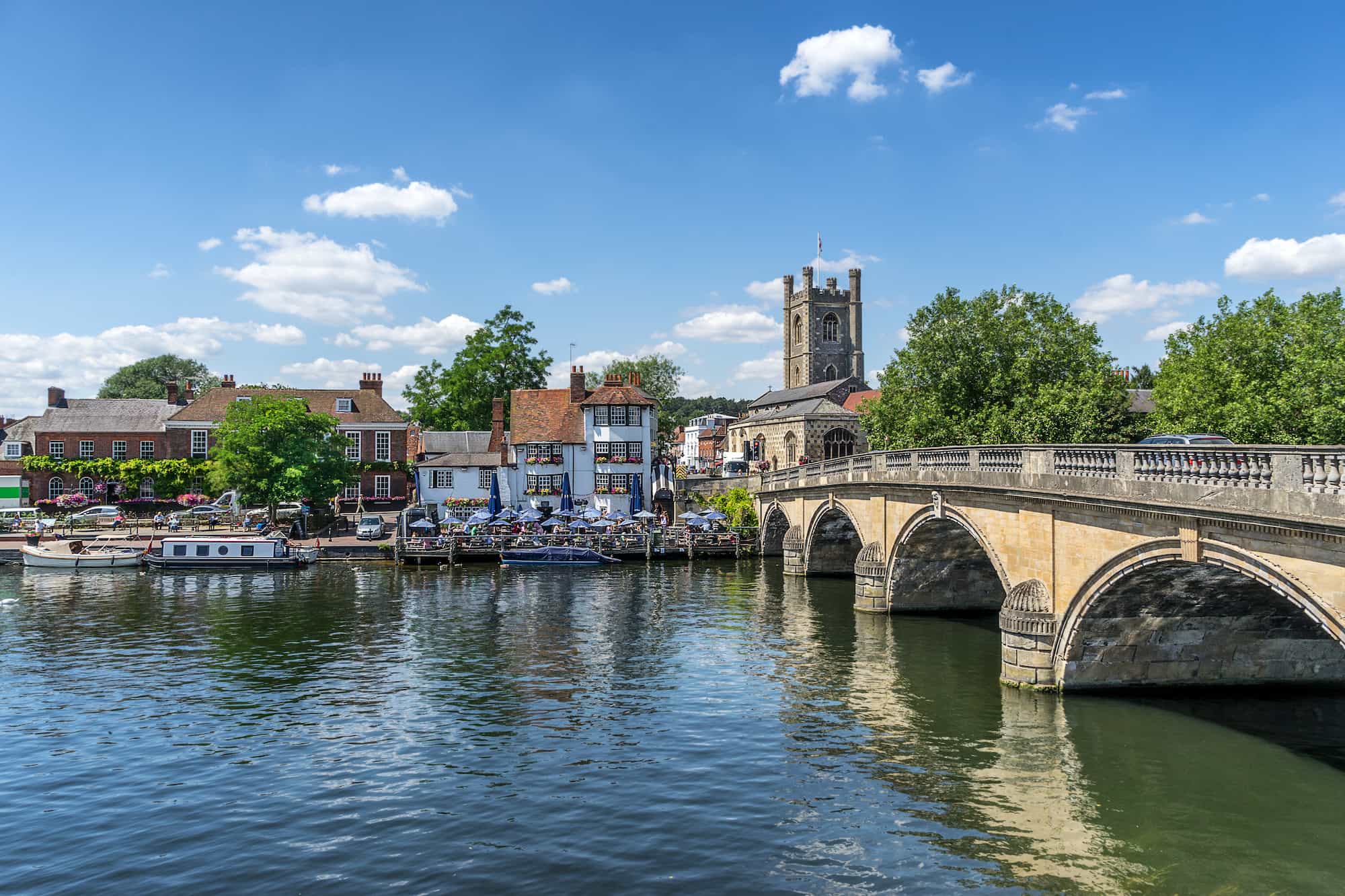 A stone bridge crosses a calm river beside historic buildings, outdoor cafes, and a church with a bell tower under a blue sky with scattered clouds.