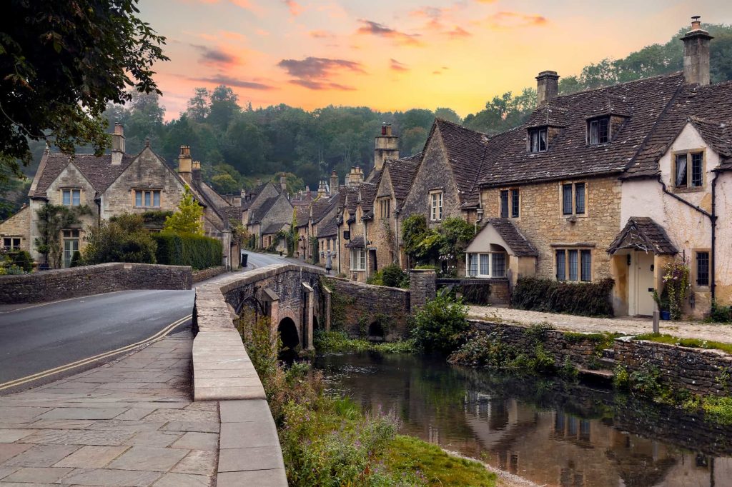 A stone bridge leads into a quaint village with old stone cottages, lush greenery, and a calm stream, all under a colorful sunset sky.