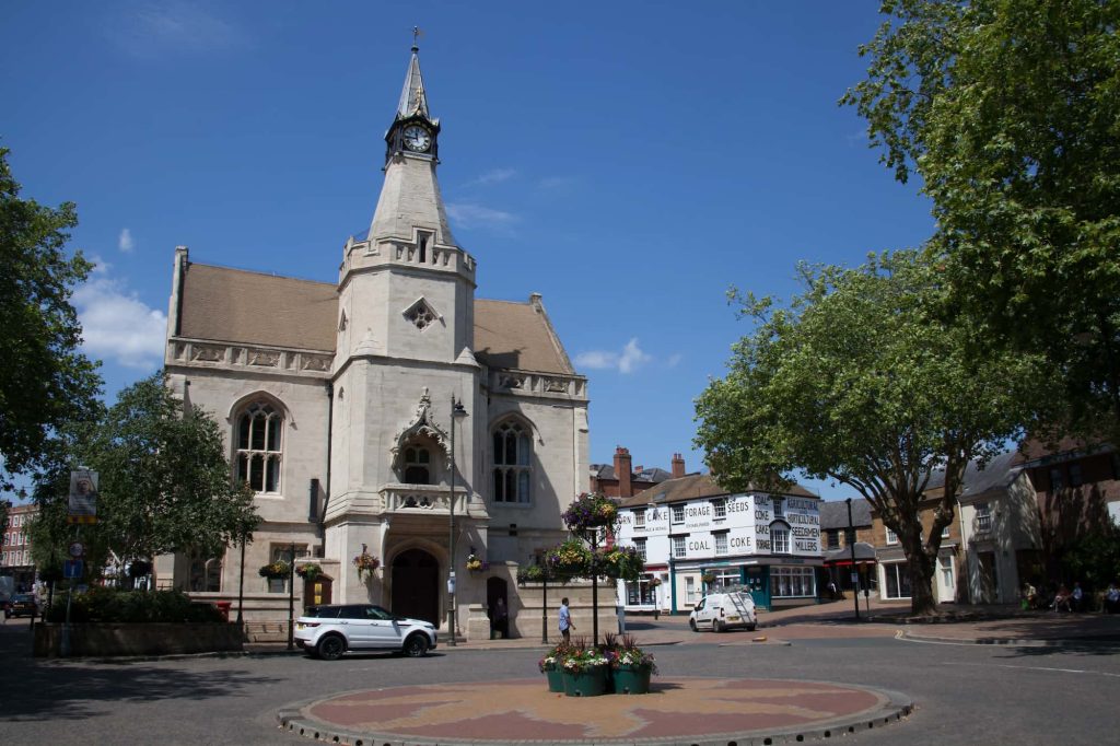A historic stone building with a clock tower stands in a sunny town square, surrounded by trees, cars, and shops. A planter with flowers is in the center foreground.
