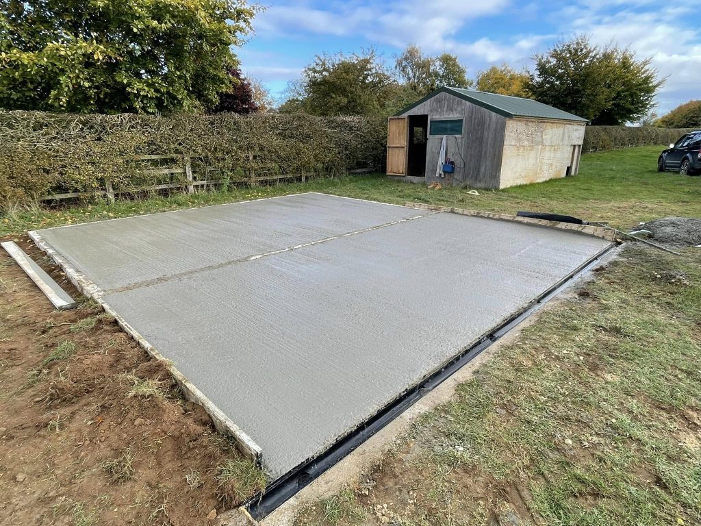 A freshly poured concrete slab is set on a grassy area near a wooden shed. Trees and a hedge border the background under a partly cloudy sky.