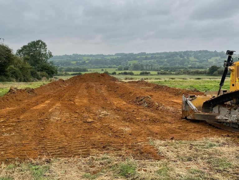A bulldozer is parked on a patch of freshly moved earth in a grassy field, with trees and rolling hills visible in the background under a cloudy sky.