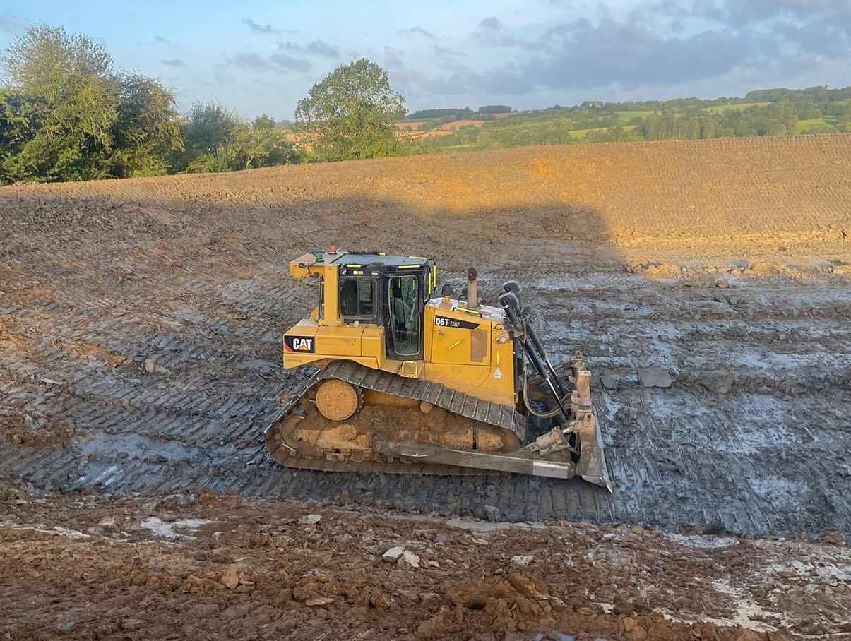 A yellow Caterpillar bulldozer moves earth on a construction site, surrounded by dirt and grassy hills under a partly cloudy sky.