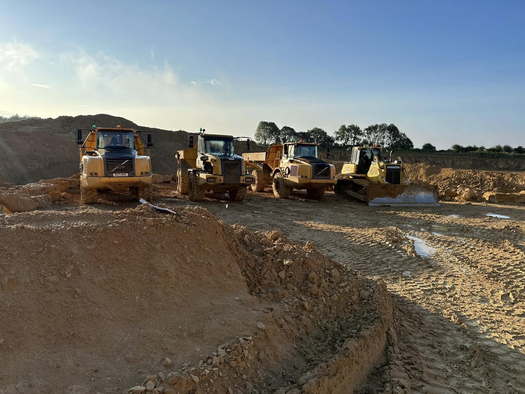 Four large yellow construction vehicles, including bulldozers and dump trucks, are parked on a dirt construction site under a clear blue sky with trees in the background.