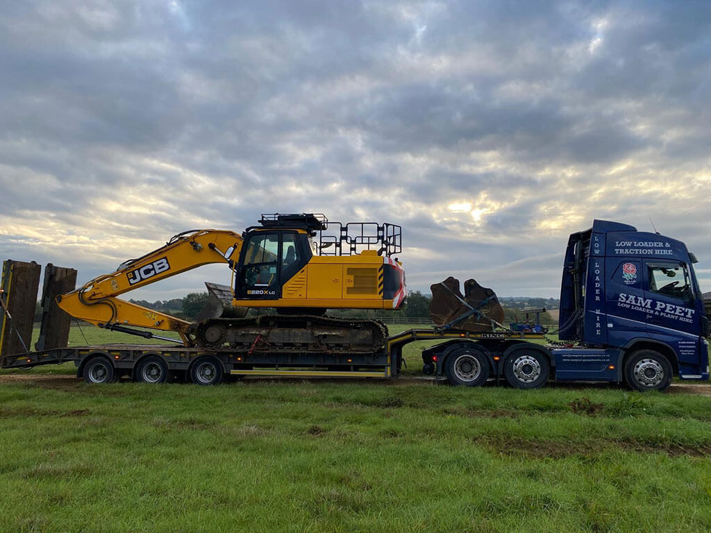 A yellow JCB excavator is loaded on a blue flatbed truck in a grassy field under a cloudy sky, with some distant trees visible in the background.