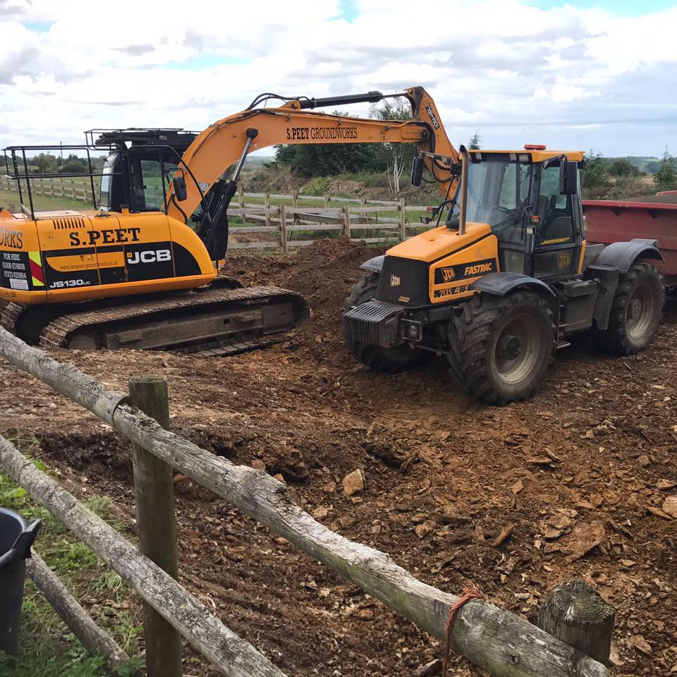 A yellow excavator and a yellow loader are working on a dirt construction site, surrounded by a wooden fence, with a field and cloudy sky in the background.
