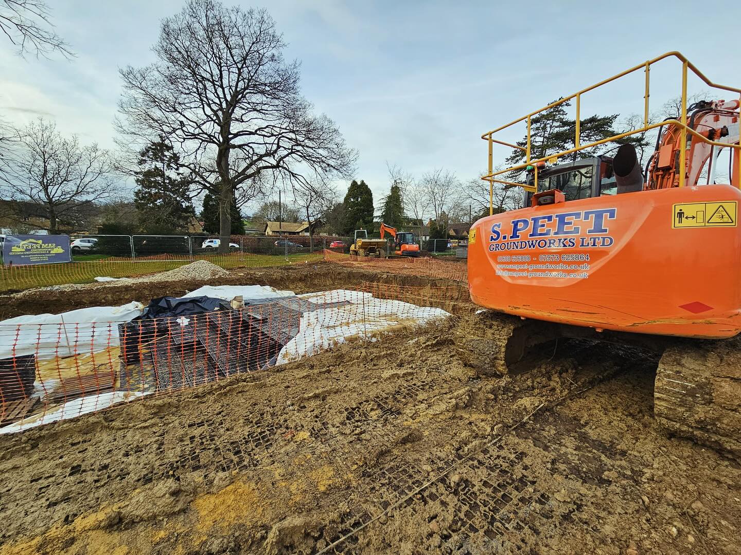 A large orange excavator labeled “S. PEET GROUNDWORKS LTD” sits on a muddy construction site with protective mats, fencing, trees, and parked cars in the background.