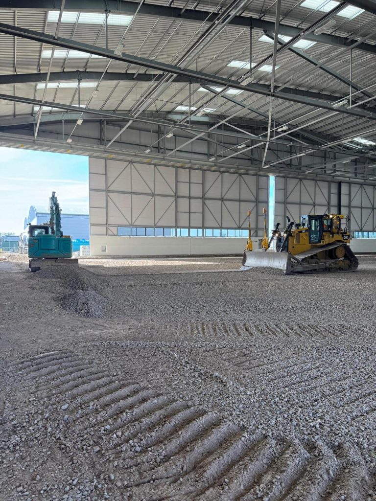 A blue excavator and a yellow bulldozer work on spreading and leveling gravel inside a large, unfinished industrial building with high ceilings and metal beams.