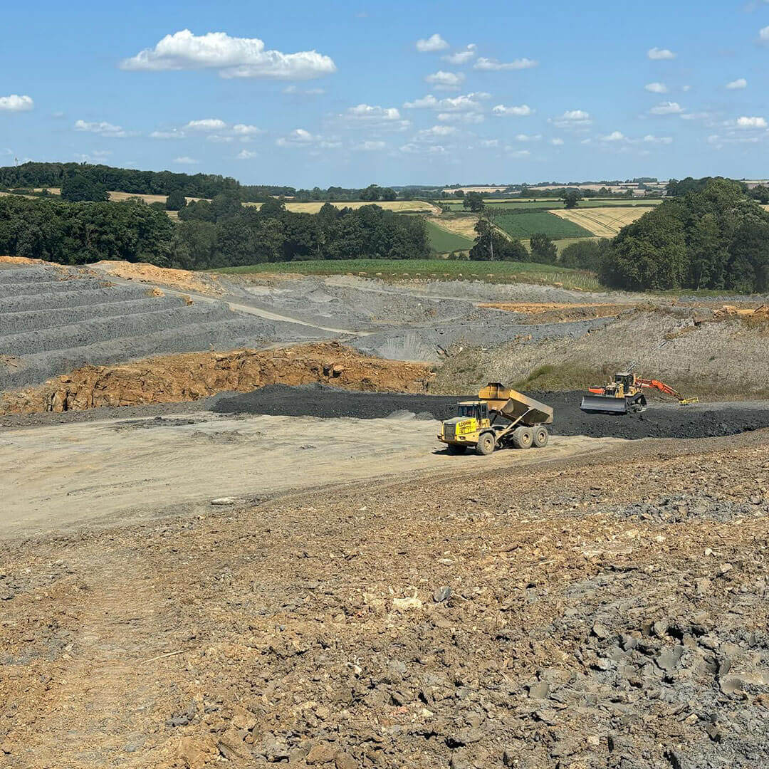 A quarry scene with a yellow dump truck and an excavator working on rocky terrain, surrounded by terraced earth, trees, and green fields under a blue sky with scattered clouds.