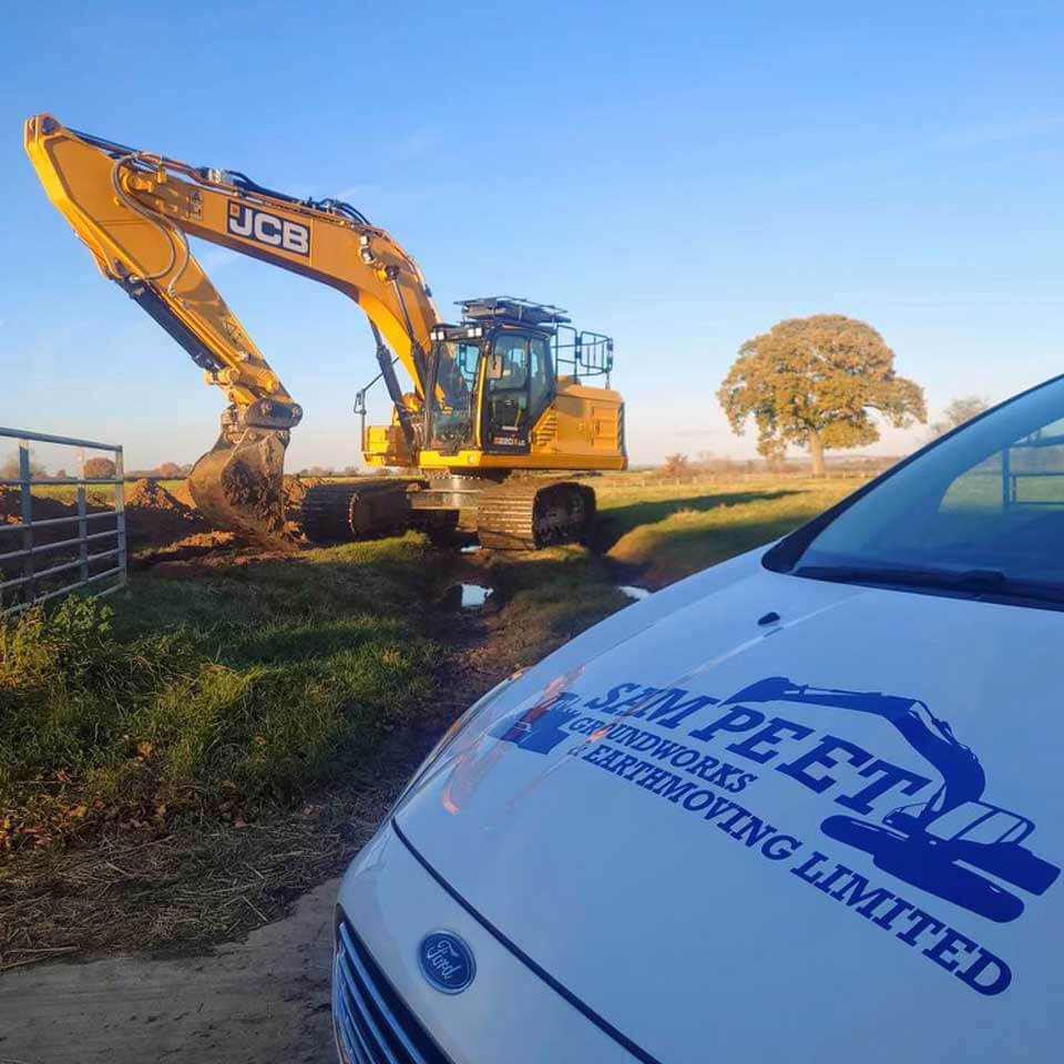 A yellow JCB excavator works on grass near a fence, with a white Simp-E-T sandworks and earthmoving limited van parked in the foreground. A tree stands in the background under a clear blue sky.