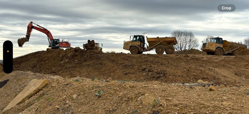 Excavator and three dump trucks work on a large dirt mound at a construction site under a cloudy sky. Trees are faintly visible in the background.