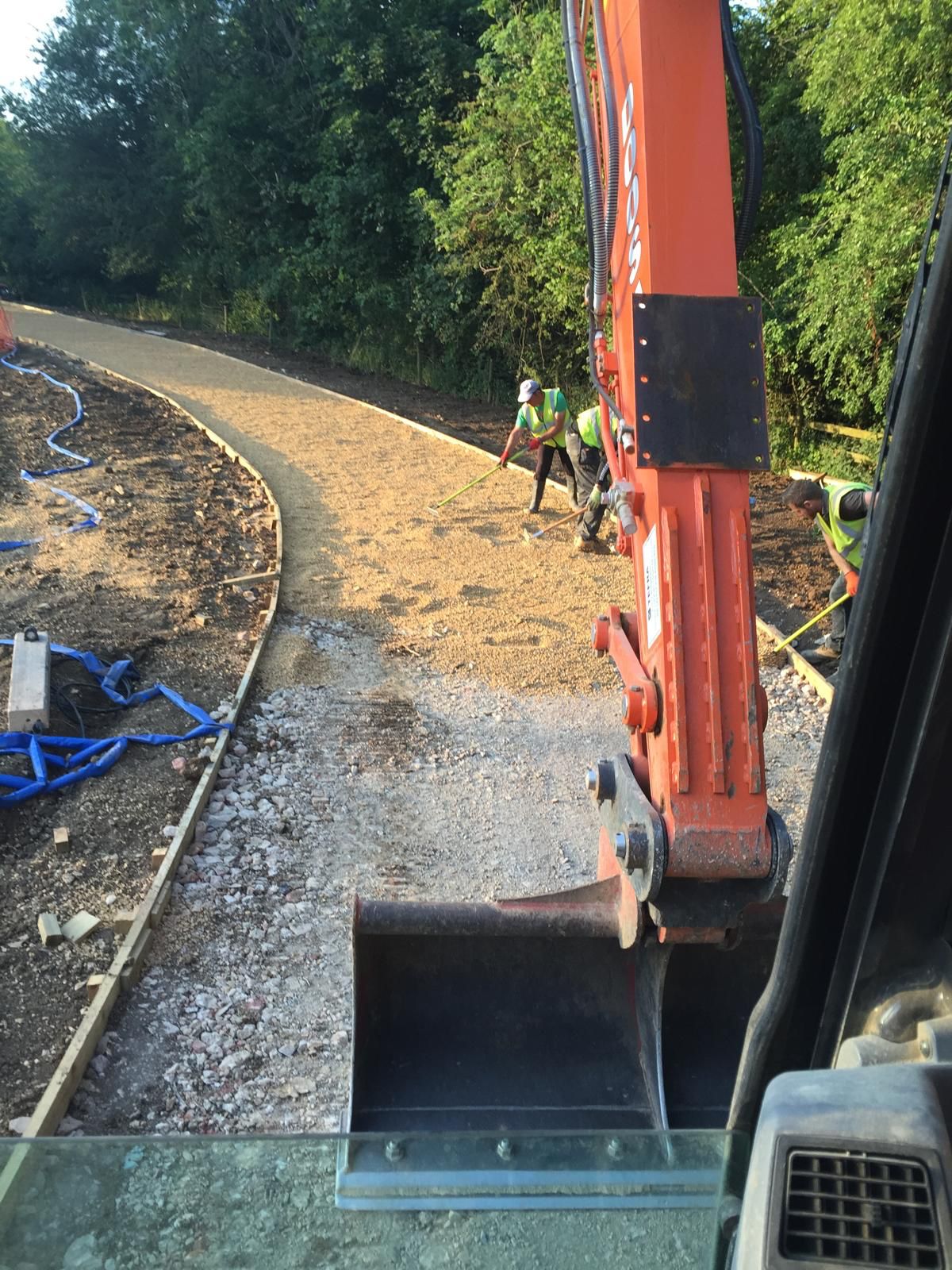Two construction workers in safety vests level gravel on a winding path, while an orange excavator is visible in the foreground. Trees line the path on both sides.