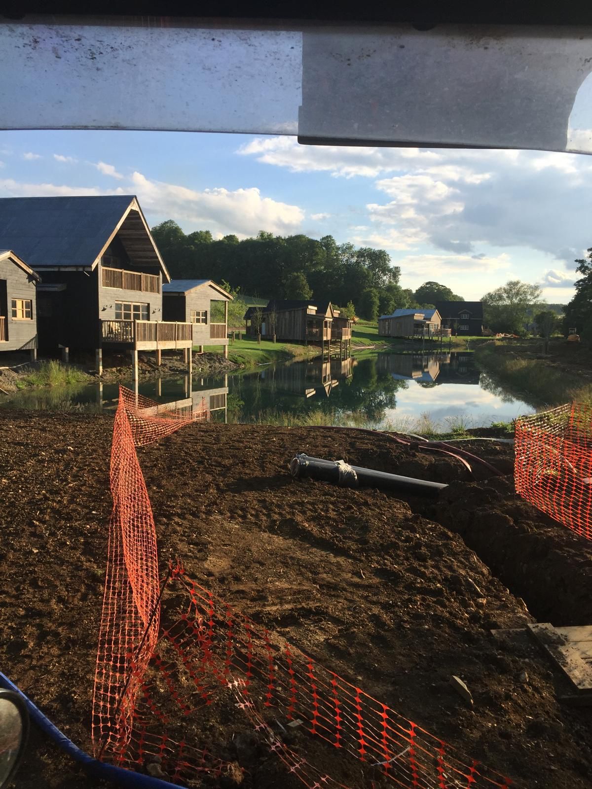 A construction site with orange fencing in the foreground and modern buildings by a calm lake in the background, reflecting trees and a partly cloudy sky.