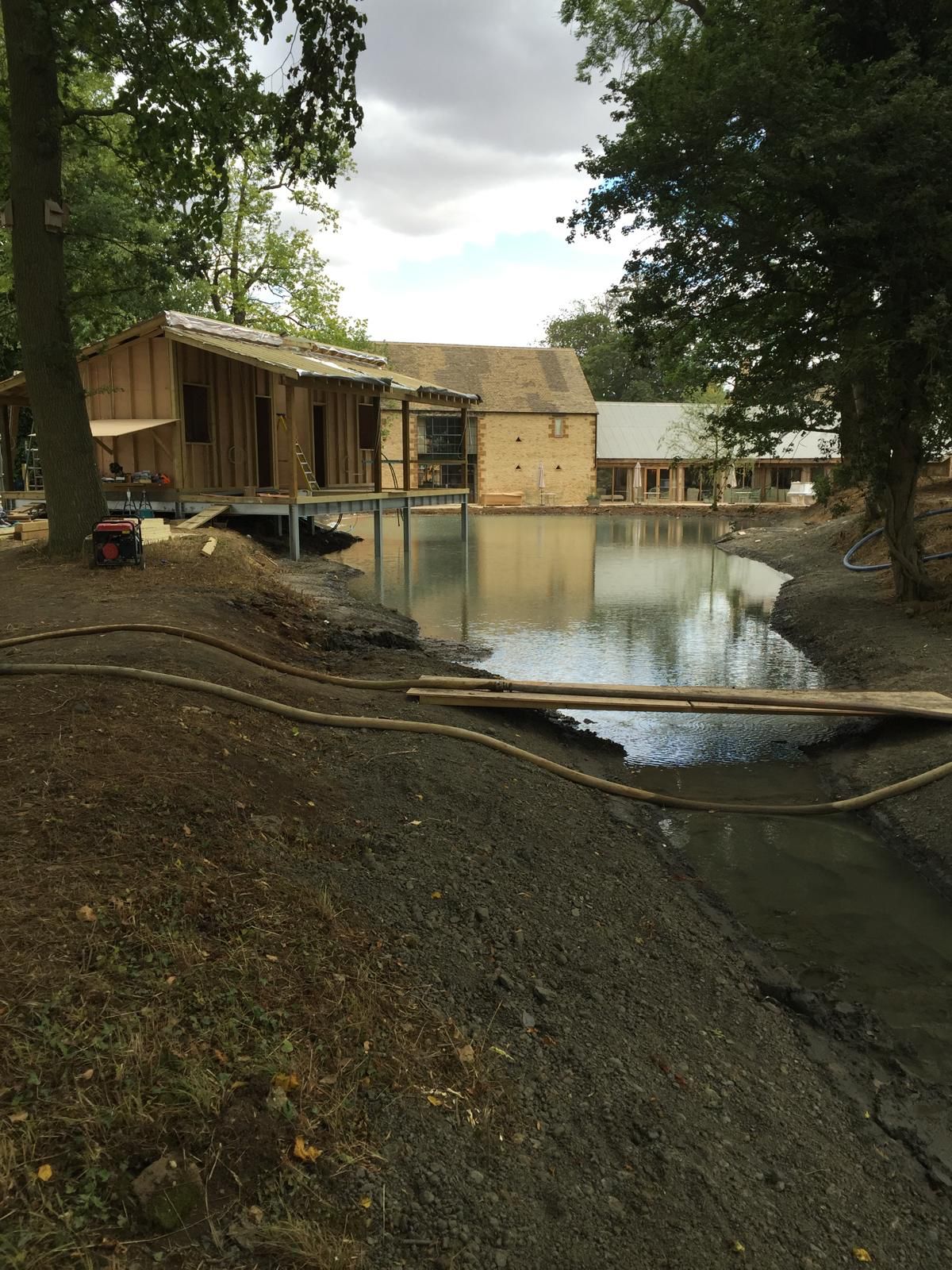 A small pond with houses under construction nearby, surrounded by trees. Building materials and hoses are visible near the water’s edge. The sky is mostly cloudy.
