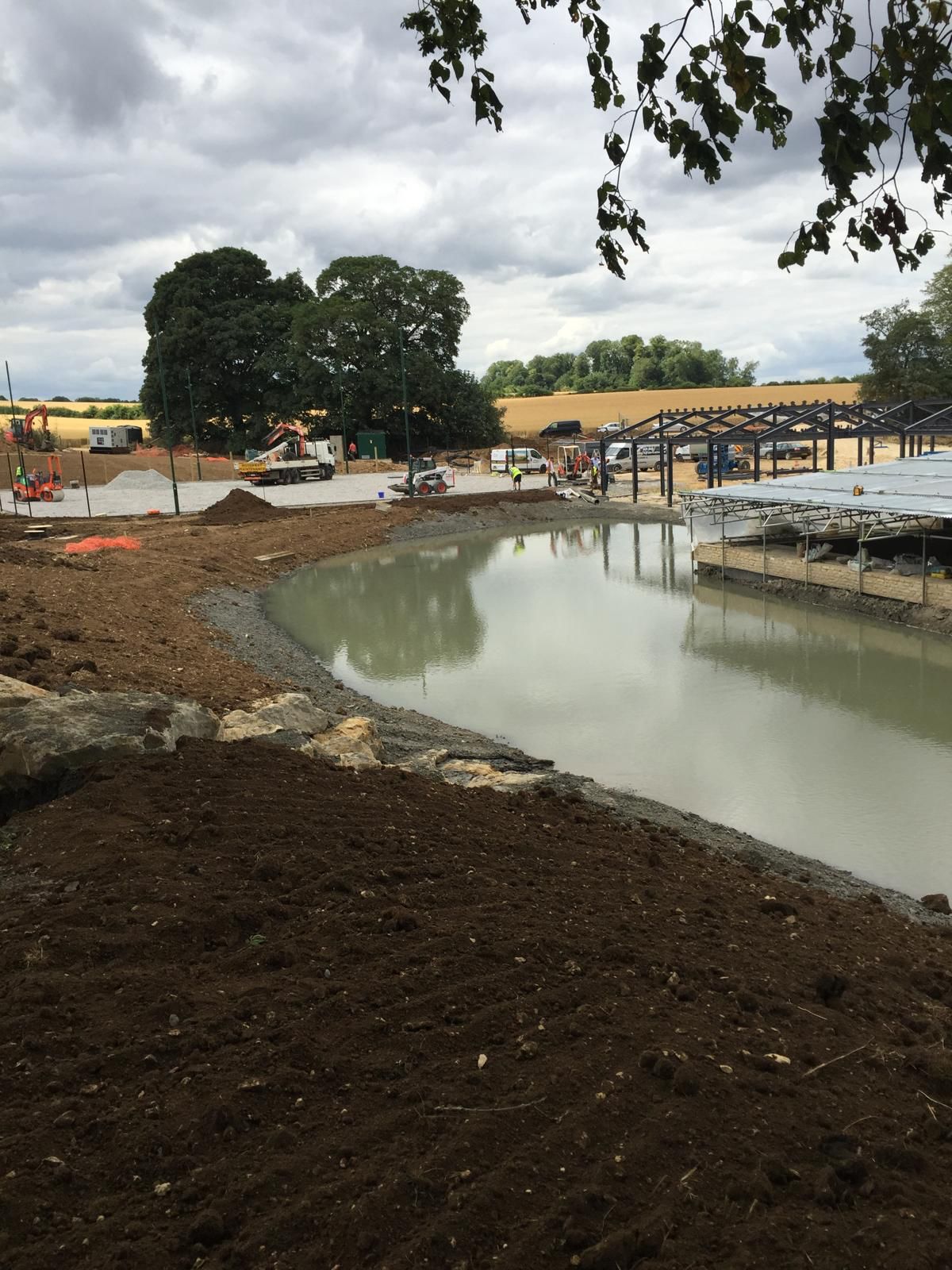 A construction site with a partially finished pond, soil around the edge, machinery, and workers in the background. Steel framework for structures is visible under a cloudy sky.
