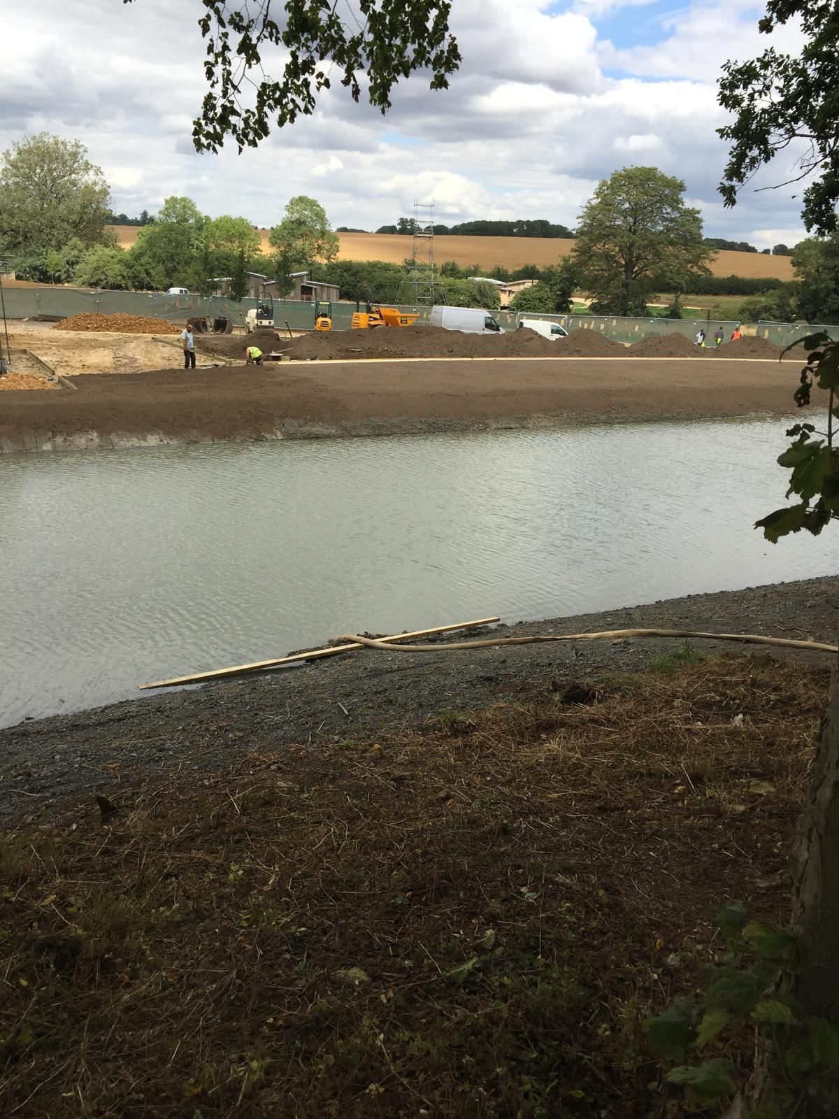 Construction workers and machinery are visible near a body of water, with grassy fields and trees in the background under a partly cloudy sky.