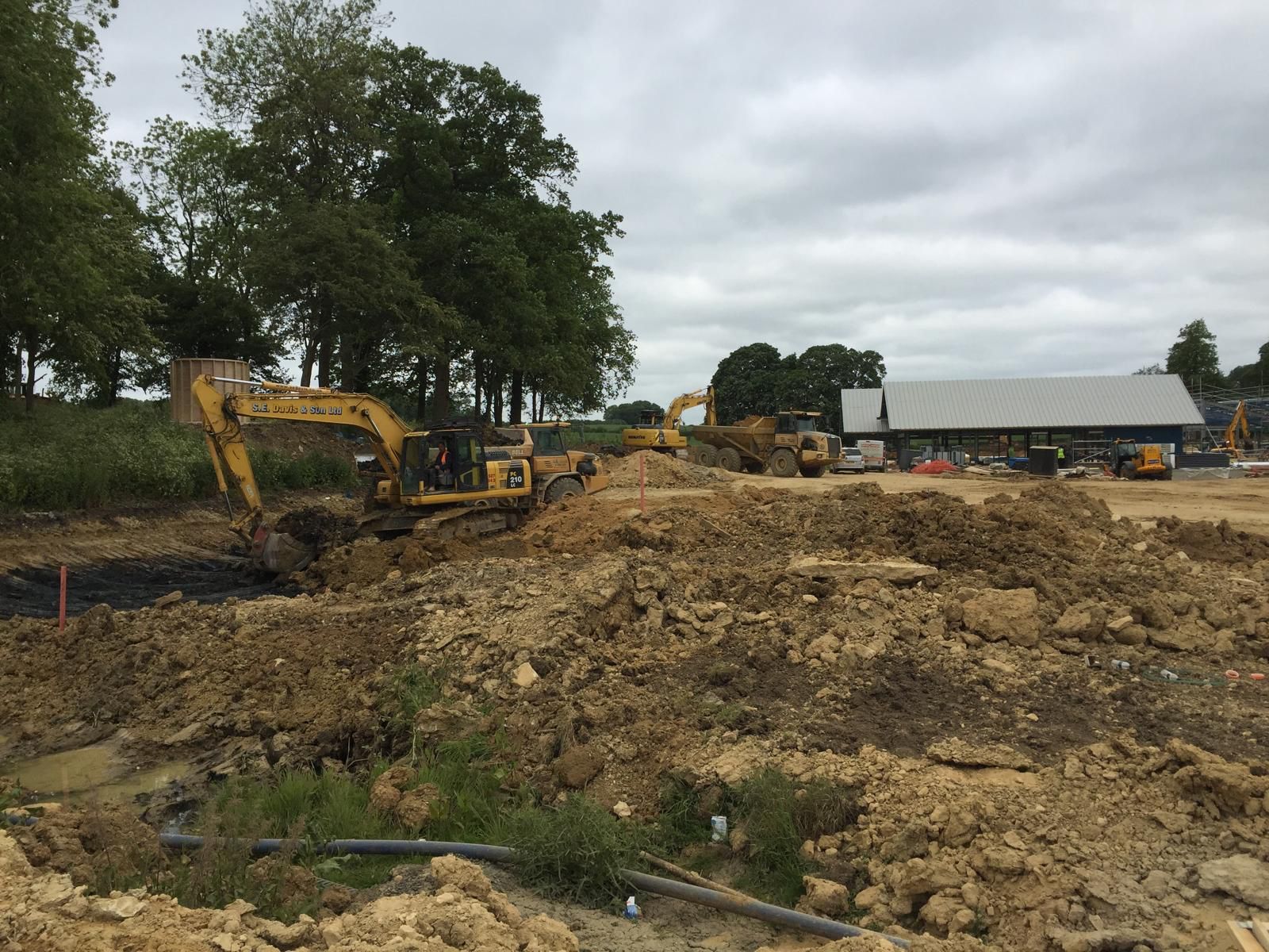 Construction site with excavators digging and moving earth; dirt piles cover the ground, with trees and a building in the background under a cloudy sky.