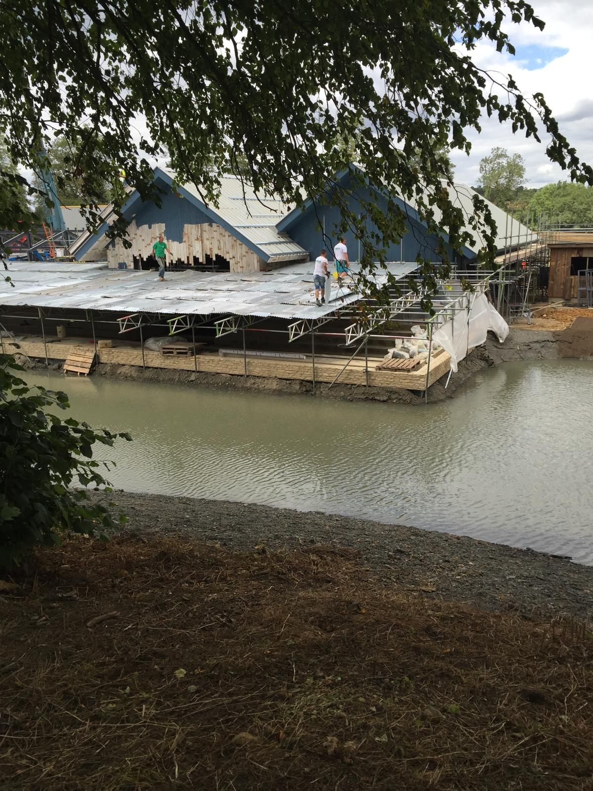 A partially constructed building with people working on a platform over water, surrounded by scaffolding and trees.