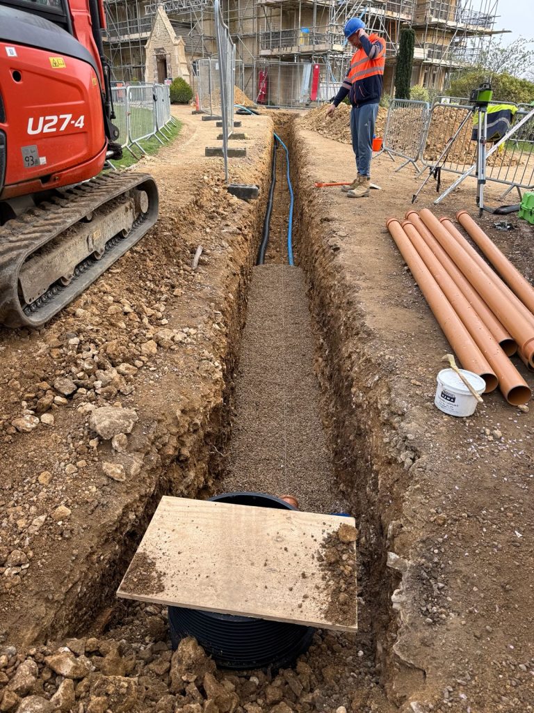 A construction worker stands beside a long, narrow trench with blue pipes, gravel, and orange pipes nearby. An excavator is on the left, and scaffolding is visible in the background.