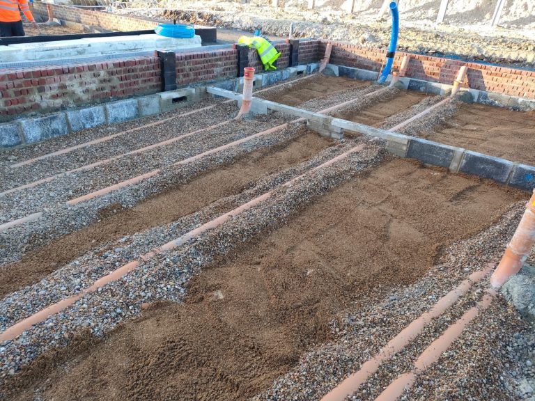 Construction site with exposed orange drainage pipes laid in gravel and sand, brick walls in the background, and a worker in a yellow vest partially visible. Blue pipes and construction materials are present.