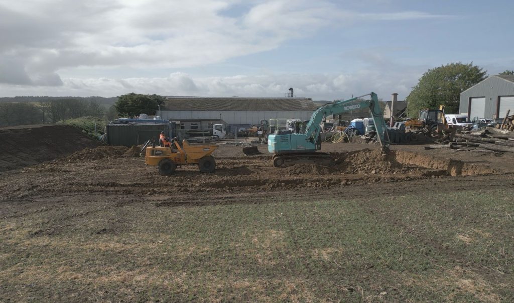 A construction site with an excavator and a small yellow dumper moving earth. Buildings, machinery, and equipment are in the background under a cloudy sky.